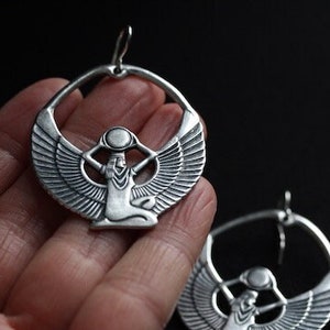 May include: Silver hoop earrings with a detailed design of a winged goddess holding a vessel on her head.
