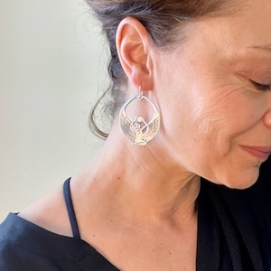 May include: Silver hoop earrings with a detailed design of a winged figure. The figure is holding a sphere in its hands.
