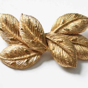 May include: A gold-colored leaf-shaped hair clip. The clip features six detailed leaves arranged in a cluster. The leaves have textured surfaces and are made of a shiny, metallic material.