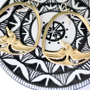 May include: A pair of gold-colored hoop earrings. Each earring features a bird design within the hoop. The earrings rest on a black and white patterned surface, creating a striking contrast. The bird design has detailed wings.