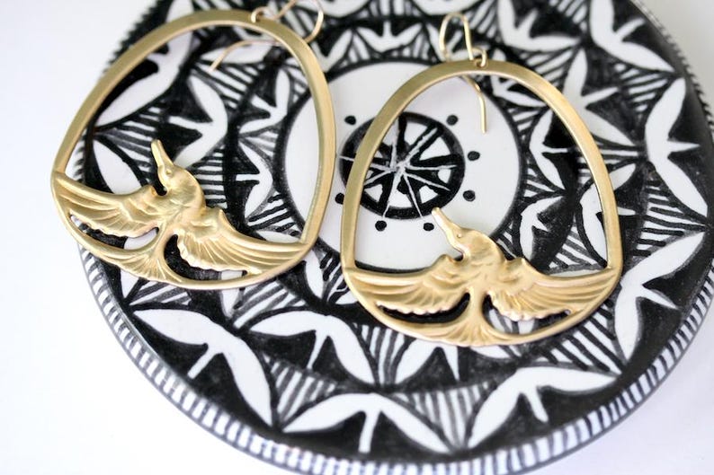 May include: A pair of gold-colored hoop earrings. Each earring features a bird design within the hoop. The earrings rest on a black and white patterned surface, creating a striking contrast. The bird design has detailed wings.