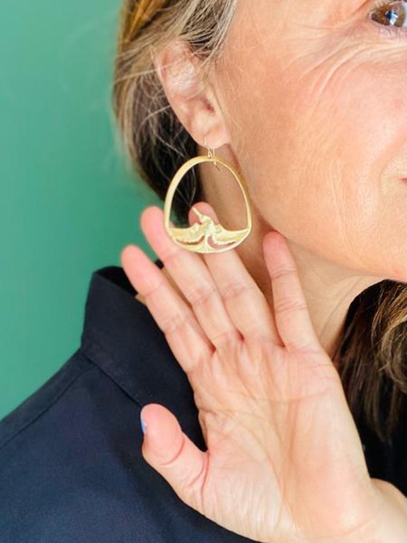 May include: Gold-toned hoop earrings with a unique bird and wave design. The earrings have a hook closure and are worn on the ear. The design is intricate and detailed, with a cutout pattern. The earrings are a stylish accessory.