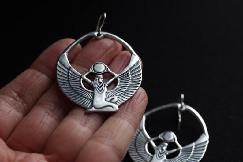 May include: Silver hoop earrings with a detailed design of a winged goddess holding a vessel on her head.