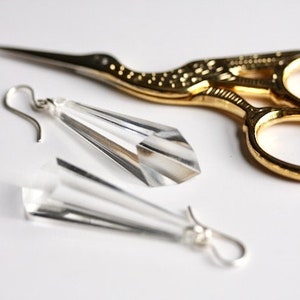 Op de afbeelding: Een paar heldere, facetgeslepen kristallen oorbellen met zilverkleurige haken, naast een goudkleurige borduurschaar. De schaar heeft een decoratief vogelontwerp op de handvatten. De items liggen op een wit oppervlak.