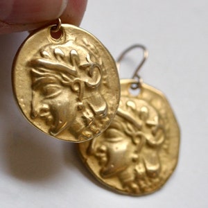 Puede incluir: Pendientes de tono dorado con un diseño de moneda redonda que presenta un perfil en relieve de la cara de una mujer. Los pendientes tienen un cierre de gancho simple.