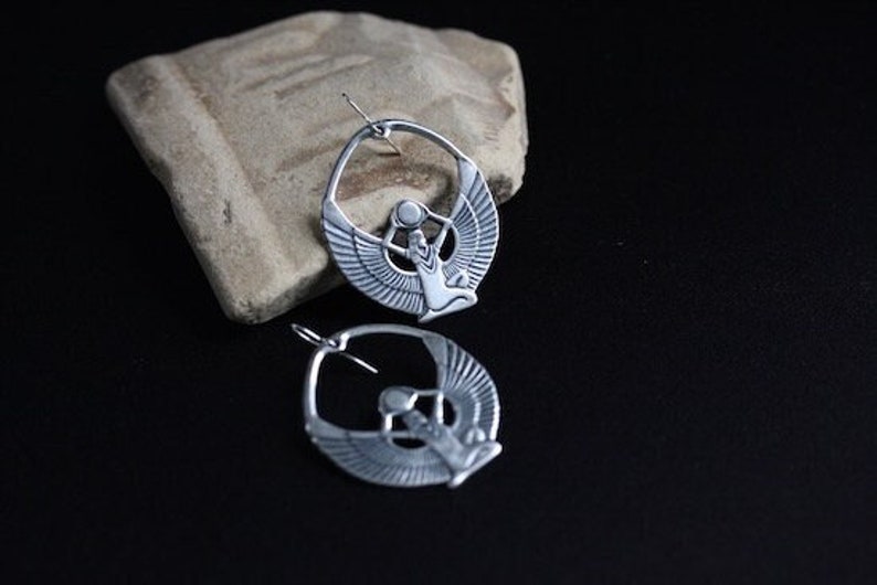May include: A pair of silver hoop earrings featuring a detailed cutout design of a winged figure holding a sphere. The earrings are hanging from a rough, textured surface.