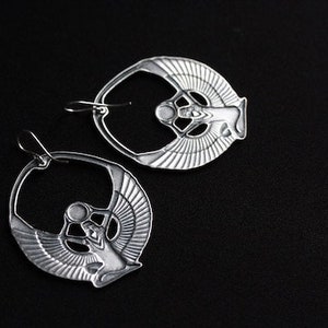 May include: A pair of silver earrings featuring a detailed design of a winged goddess figure. The earrings are hoop-shaped and have a polished finish.