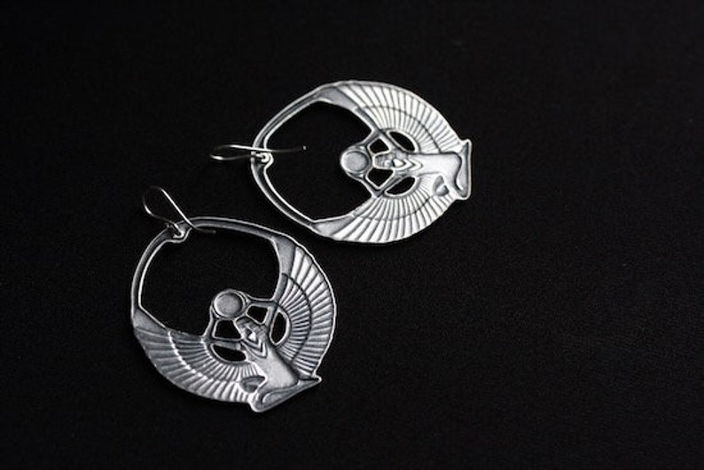 May include: A pair of silver earrings featuring a detailed design of a winged goddess figure. The earrings are hoop-shaped and have a polished finish.