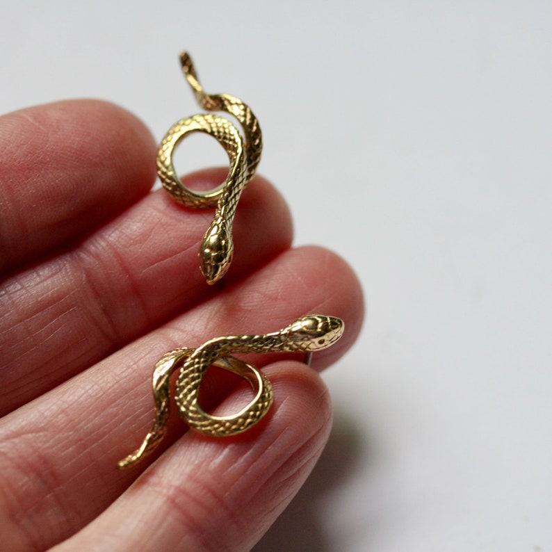 May include: Two gold-colored snake earrings. The snakes are curled into a circle and have detailed scales.