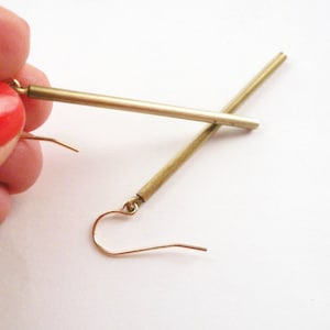 May include: A pair of gold-toned earrings with a long, thin bar design. The earrings are made of metal and have a simple, minimalist style.