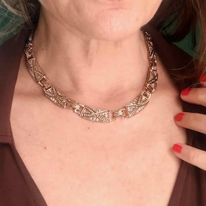 May include: A gold-toned necklace with an intricate geometric design. The necklace is composed of rectangular and patterned links, creating a bold statement piece. The necklace is worn around the neck.