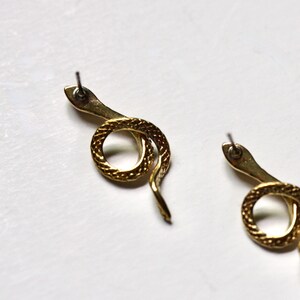 May include: A pair of gold snake earrings with a textured finish. The earrings are in the shape of a snake curled into a circle.