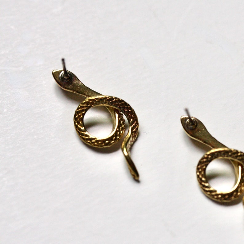 May include: A pair of gold snake earrings with a textured finish. The earrings are in the shape of a snake curled into a circle.