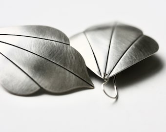 May include: A pair of silver-toned earrings shaped like stylized leaves with a brushed metal finish. Each earring features distinct etched lines radiating from the base to the pointed tip, with a simple curved wire hook attached to the back.