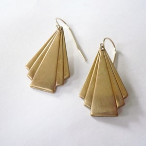 May include: A pair of gold-toned earrings with a geometric, fan-shaped design. The earrings are made of metal and have a simple, elegant style.