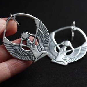 May include: A pair of silver hoop earrings with a detailed design of a winged goddess holding a sphere. The earrings are large and have a circular shape.