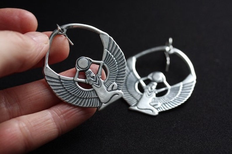 May include: A pair of silver hoop earrings with a detailed design of a winged goddess holding a sphere. The earrings are large and have a circular shape.