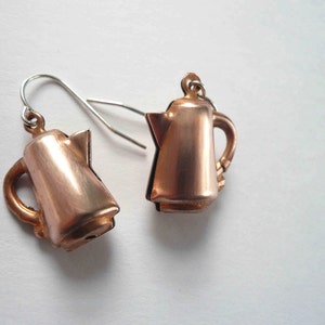 May include: A pair of copper-colored earrings shaped like coffee pots. The earrings have silver hooks.