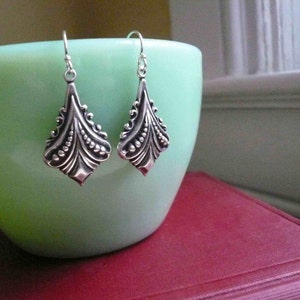 May include: A pair of silver dangle earrings with a detailed, ornate design. The earrings are shaped like a teardrop with a scalloped edge and a textured surface.