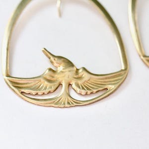 May include: Gold-toned metal earrings featuring a bird design. The earrings have a curved top and a detailed bird in the center with outstretched wings. The bird is in flight, adding a touch of nature-inspired elegance to the jewelry.