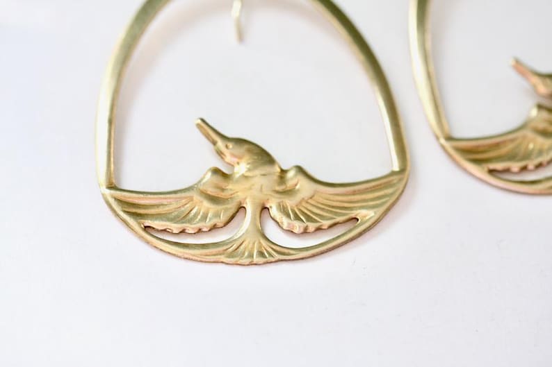 May include: Gold-toned metal earrings featuring a bird design. The earrings have a curved top and a detailed bird in the center with outstretched wings. The bird is in flight, adding a touch of nature-inspired elegance to the jewelry.