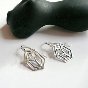 Könnte beinhalten: Ein Paar silberne Ohrringe mit geometrischem Design. Die Ohrringe sind sechseckig mit einem stilisierten Blumenmuster im Inneren.