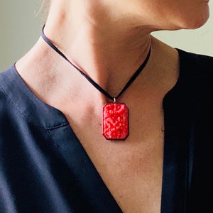 May include: A red carved pendant necklace with a black cord. The pendant is rectangular and features a floral design.
