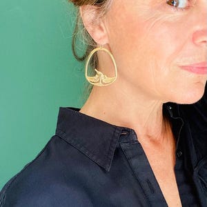 May include: Gold-toned, open-frame earrings with a bird design. The earrings are oval-shaped and feature a detailed bird silhouette within the frame. The background is a solid teal color, and the person is wearing a black collared shirt.