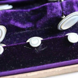 May include: A set of vintage cufflinks and shirt studs displayed in a purple velvet-lined box. The cufflinks have an oval mother-of-pearl center with a silver-tone border. The shirt studs are round with a similar design. The box is a light brown color.