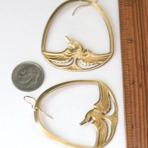 May include: A pair of gold-colored earrings. Each earring features a bird design within an oval frame. A ruler shows measurements in inches. A dime is next to the earrings for size comparison.