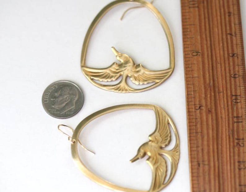 May include: A pair of gold-colored earrings. Each earring features a bird design within an oval frame. A ruler shows measurements in inches. A dime is next to the earrings for size comparison.