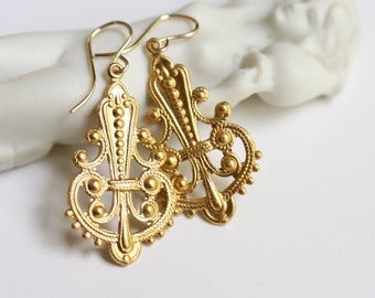 May include: A pair of ornate gold-colored earrings with a filigree design. The earrings feature a decorative, symmetrical pattern with small, raised details and a hook closure. The earrings are displayed against a white background.
