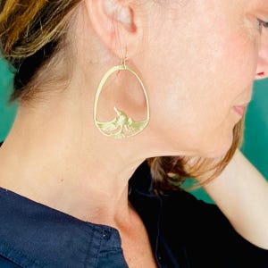 May include: Gold-toned bird earrings. The earrings feature a bird design within an oval frame. The bird is depicted with outstretched wings. The earrings are hanging from a gold-colored hook.