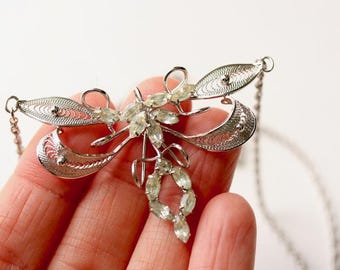 May include: Silver-toned filigree brooch with a delicate, openwork design. The brooch features a central cluster of clear, oval-shaped stones and a chain. The wings have a detailed, lacy pattern, creating an elegant, vintage aesthetic.