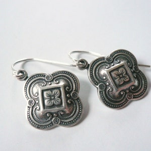 Puede incluir: Un par de pendientes de plata con un diseño cuadrado que presenta un motivo floral. Los pendientes tienen un diseño delicado e intrincado.