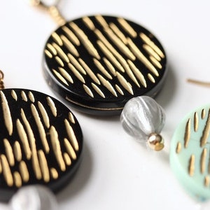 May include: A pair of dangle earrings with black and green round ceramic discs with gold accents. Each earring has a silver bead.