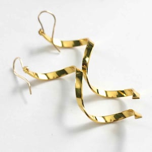 Puede incluir: Un par de pendientes dorados con forma de cinta. Los pendientes tienen un diseño en espiral con cierre de gancho. Los pendientes metálicos brillantes se muestran sobre un fondo blanco. Los pendientes son una joya.