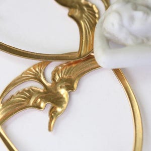 May include: Close-up of a gold-colored, Art Nouveau-style decorative element. The design features stylized bird and leaf motifs, with a white porcelain figure partially visible. The piece has a delicate, ornate appearance.