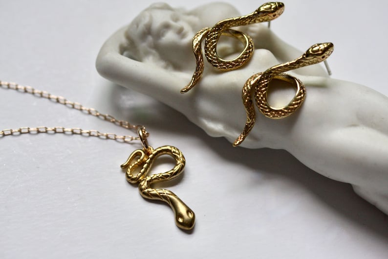 May include: A gold snake necklace and earrings set. The necklace features a delicate chain and a pendant of a coiled snake. The earrings are simple studs with a snake design.