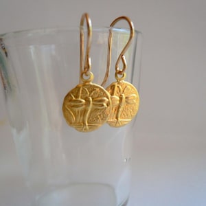 May include: Gold dangle earrings with a dragonfly design. The earrings are hanging from a glass.