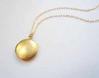 Può includere: Un ciondolo a medaglione rotondo in oro con finitura metallica spazzolata, sospeso a una delicata catenina d'oro con maglie rettangolari. Il medaglione presenta una superficie circolare liscia ed è fissato alla catena da un piccolo anello.