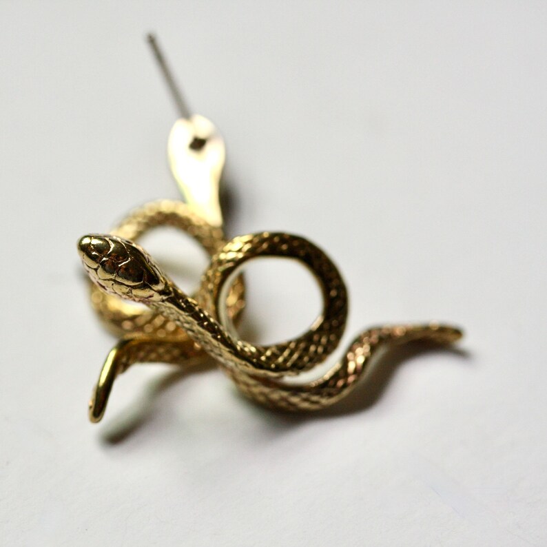 May include: Gold-toned snake earring with a textured surface. The snake is coiled and has a small head with a pointed nose.