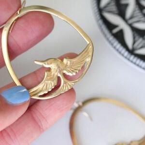 May include: Gold-toned bird earrings. Each earring features a bird design within a circular frame. The earrings are held up against a white surface, with a black and white patterned plate in the background.