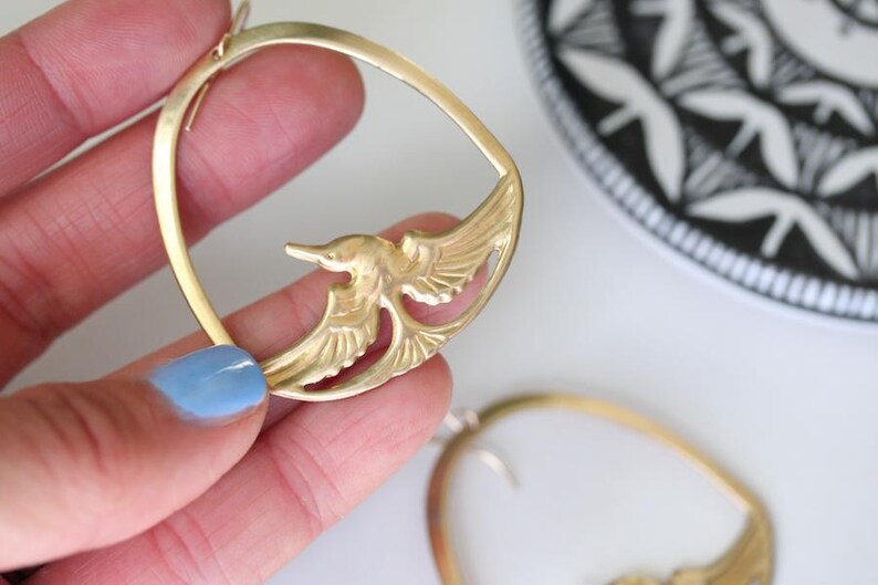 May include: Gold-toned bird earrings. Each earring features a bird design within a circular frame. The earrings are held up against a white surface, with a black and white patterned plate in the background.