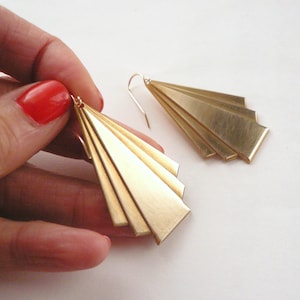 May include: A pair of gold-toned earrings with a geometric, fan-shaped design. Each earring is made of multiple thin, overlapping pieces of metal.