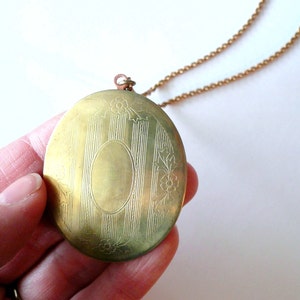 May include: A vintage gold-toned locket necklace. The oval locket features vertical lines and floral accents. The locket is suspended from a delicate chain. The locket is held in a hand.