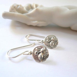 May include: A pair of silver dangle earrings with a bee design. The bee is embossed on a round disc.