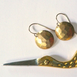 May include: A pair of round, gold-toned earrings with a hammered, textured surface. Each earring features a curved wire hook for attachment. Positioned near the earrings is a gold-colored metal tool shaped like a bird with a pointed beak.