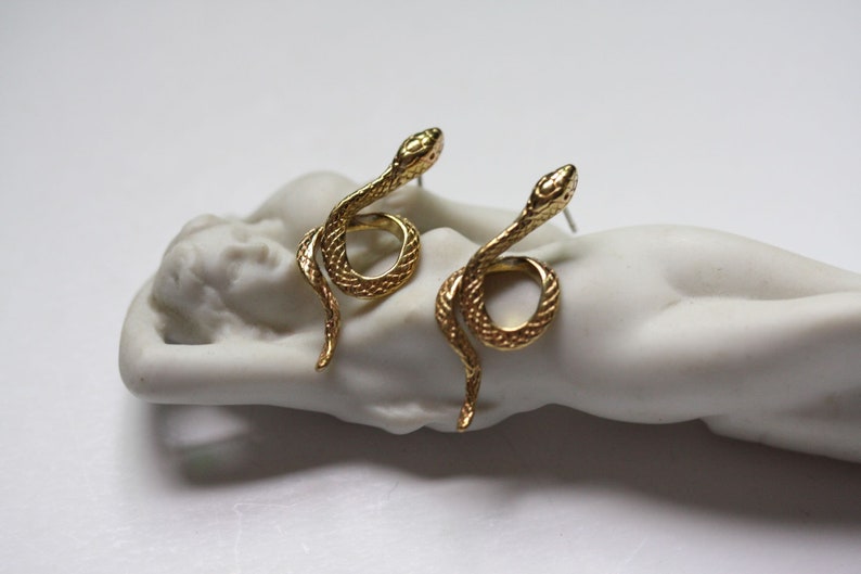 May include: A pair of gold snake earrings. The snakes are curled up with their heads facing forward. The earrings are on a white background.