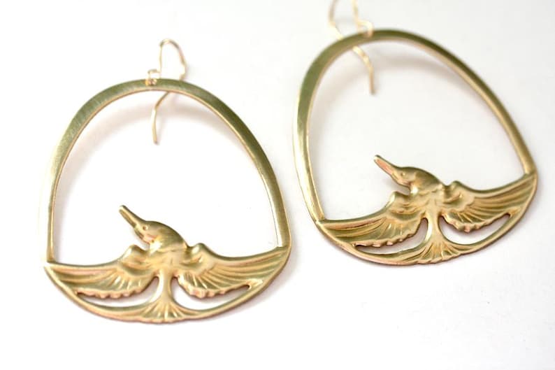 May include: A pair of gold-colored bird earrings. Each earring features a stylized bird design within an arched frame. The birds have outstretched wings and are facing upwards. The earrings are designed to hang from the ear.